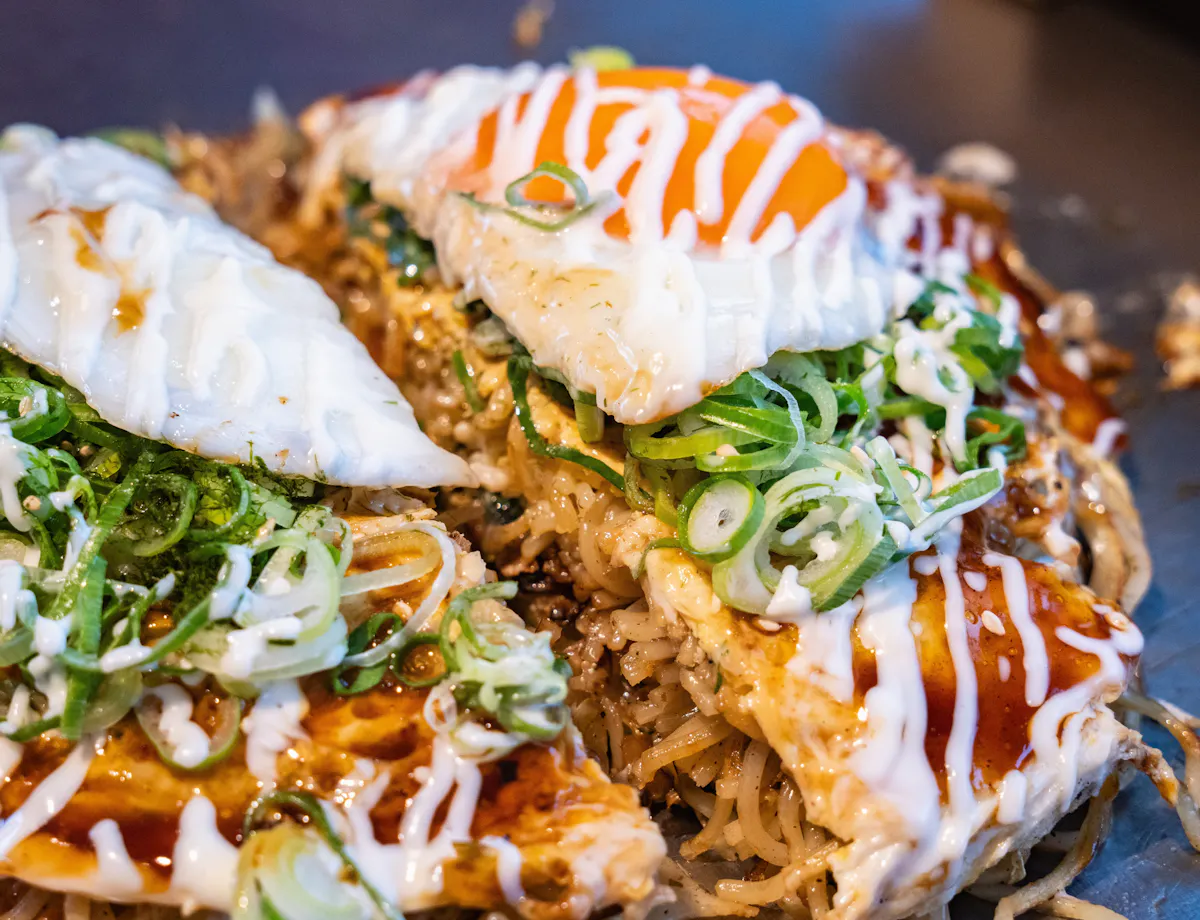 Okonomiyaki Close-up of Japanese okonomiyaki topped with fried eggs, sliced green onions, mayonnaise, and savory sauce, showing vibrant colors and textures of the ingredients.