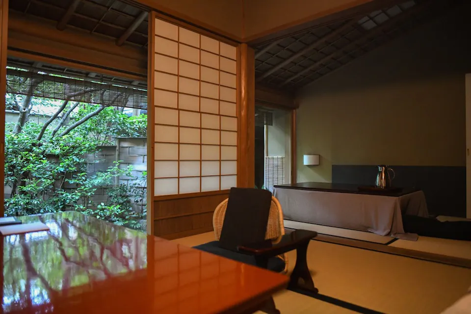 一个传统的日本房间，配有滑动的木质拉门，榻榻米地板和极简主义家具。一张低矮的桌子上放着一个水壶，靠近地上的一个垫子。外面，一座宁静的花园，郁郁葱葱的绿色植物透过敞开的门可见。