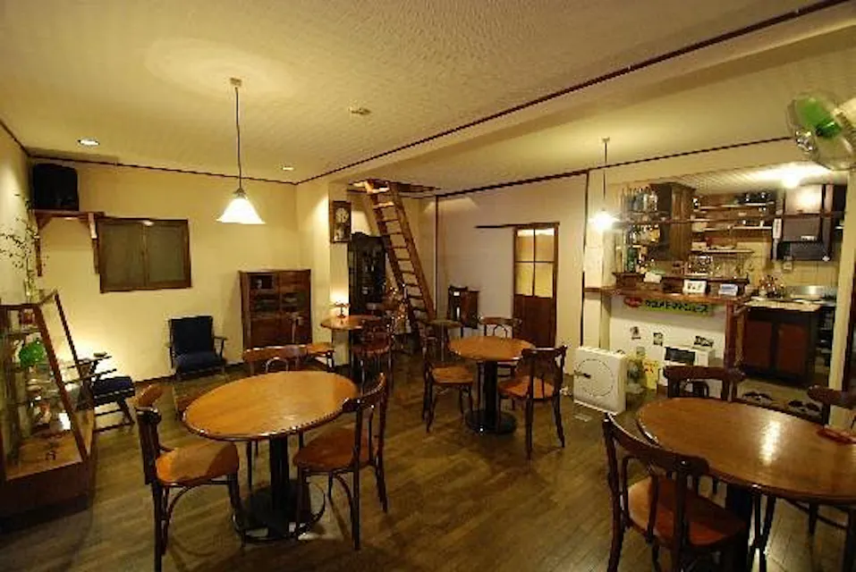 A cozy cafe interior with wooden furniture, round tables, and chairs. The room features warm lighting, shelves with decorations, and a counter area. A wooden staircase leads to an upper level, and there are various small decorative items creating a homely atmosphere.