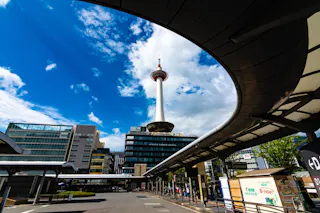 A clear day shows a modern cityscape with a prominent communications tower at its center, featuring a circular observation deck near the top. Surrounding the tower are contemporary buildings, and several covered walkways lead to the bustling urban area.