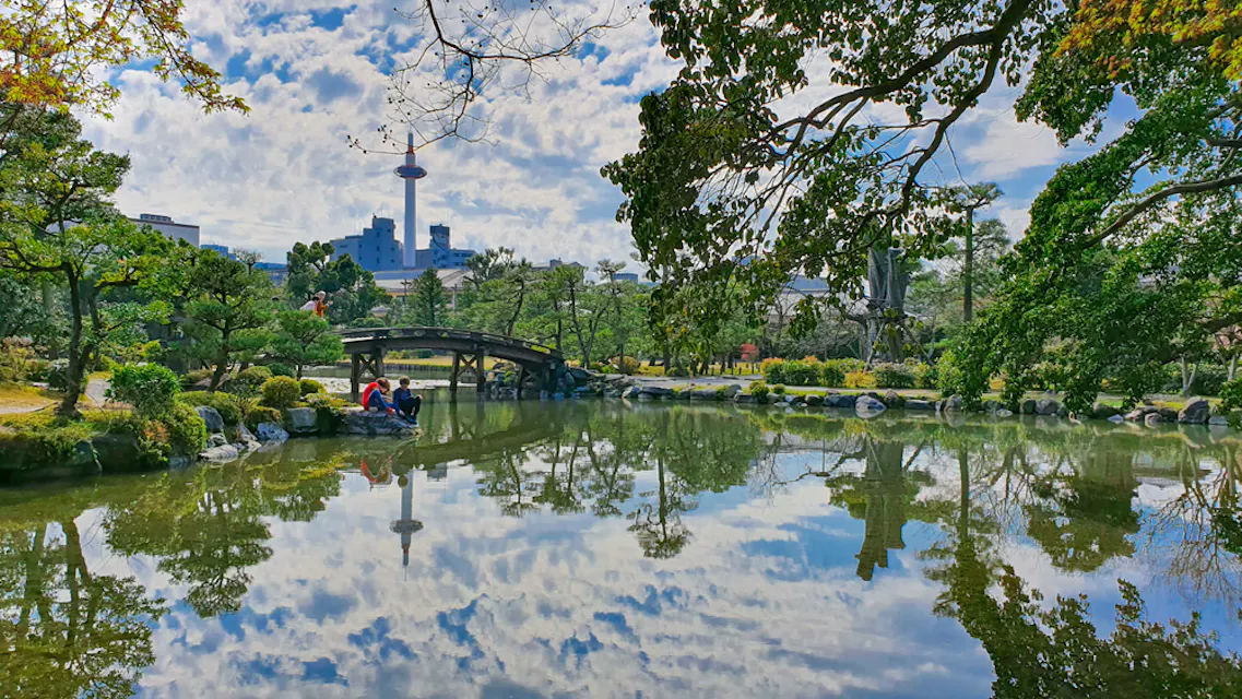 A tranquil Japanese garden features a serene pond reflecting a cloudy sky, lush trees, and a small arched bridge. In the distance, a cityscape with a tall tower is visible. Two people, one with a red umbrella, stand near the pond's edge.