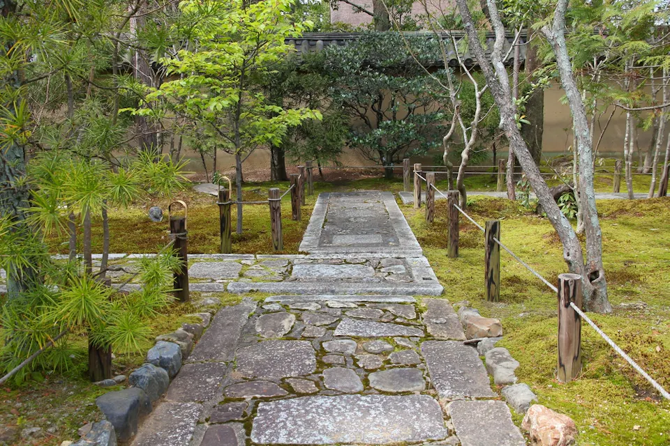 A serene Japanese garden with a moss-covered ground, stone paths, and wooden posts connected by ropes. The garden is filled with various trees and shrubs, creating a tranquil, natural atmosphere. In the background, there's a traditional wall partially visible.