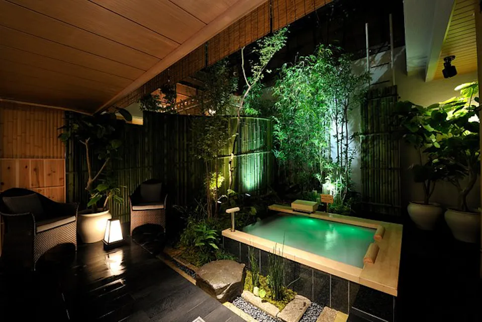 A cozy indoor-outdoor spa area featuring a small hot tub surrounded by lush greenery and bamboo. The space is softly lit, with wicker chairs and lanterns adding to the serene ambiance. The ceiling is wooden, and the floor is black tile.