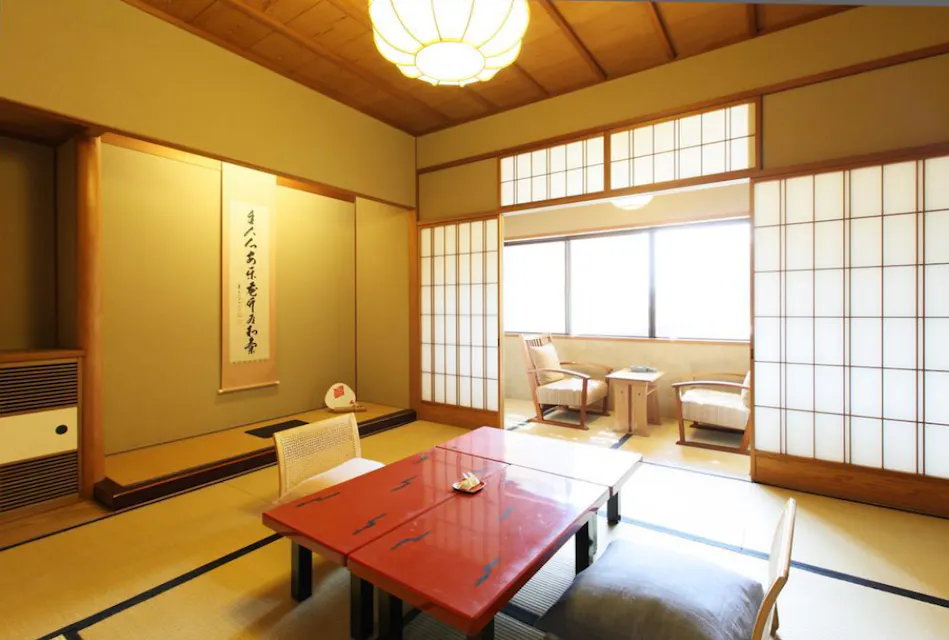 A traditional Japanese room with tatami flooring, a red lacquered low table, and floor cushions. Shōji sliding doors lead to a bright sitting area with two chairs and a small table by a window. A hanging scroll decorates one of the walls next to a small alcove.
