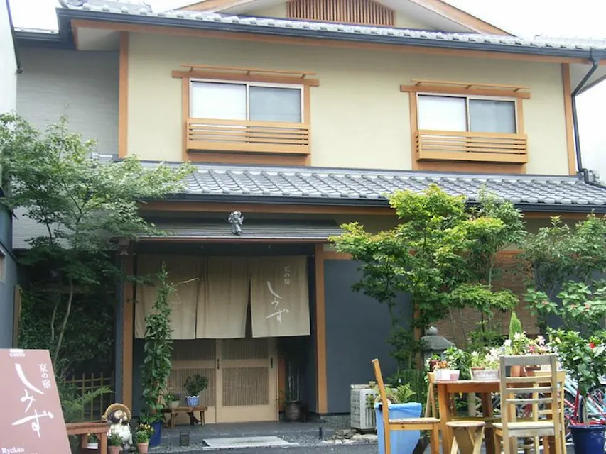 A traditional Japanese-style building with beige walls, wooden trims, and a tiled roof. The entrance features a fabric awning with Japanese characters. Outside, there are potted plants, a small table with chairs, and a sign with more Japanese text.