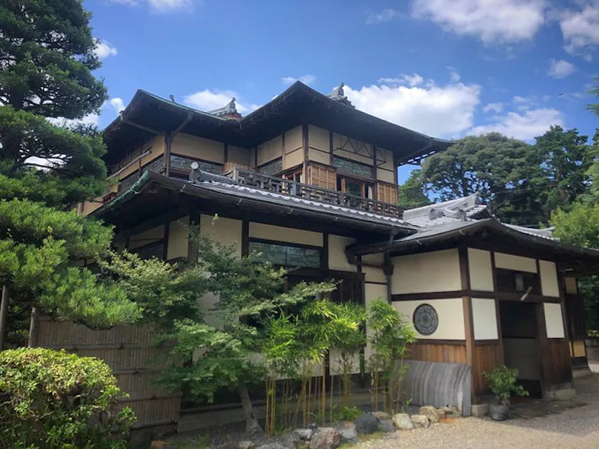 一座传统的日本建筑，具有木质装饰和瓦屋顶，被郁郁葱葱的绿树环绕。天空清澈，几朵白云点缀其间，一条石头小径通往入口。