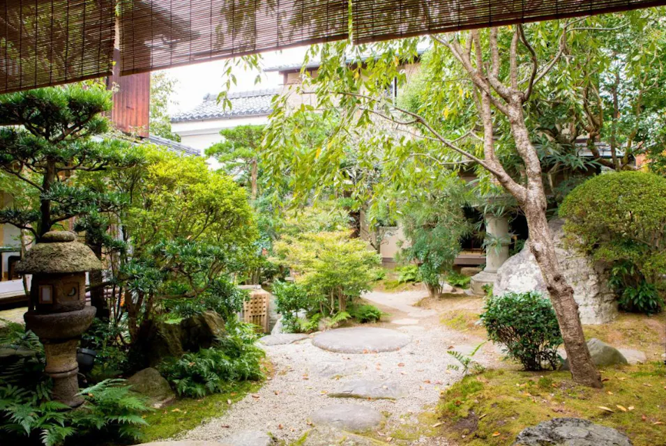 一个宁静的日本花园，规划精美，有郁郁葱葱的绿色植物、石灯、修剪整齐的灌木和石头小径。背景中部分可见一座传统的木制建筑，周围被悬挂的竹帘和繁茂的树木框住。