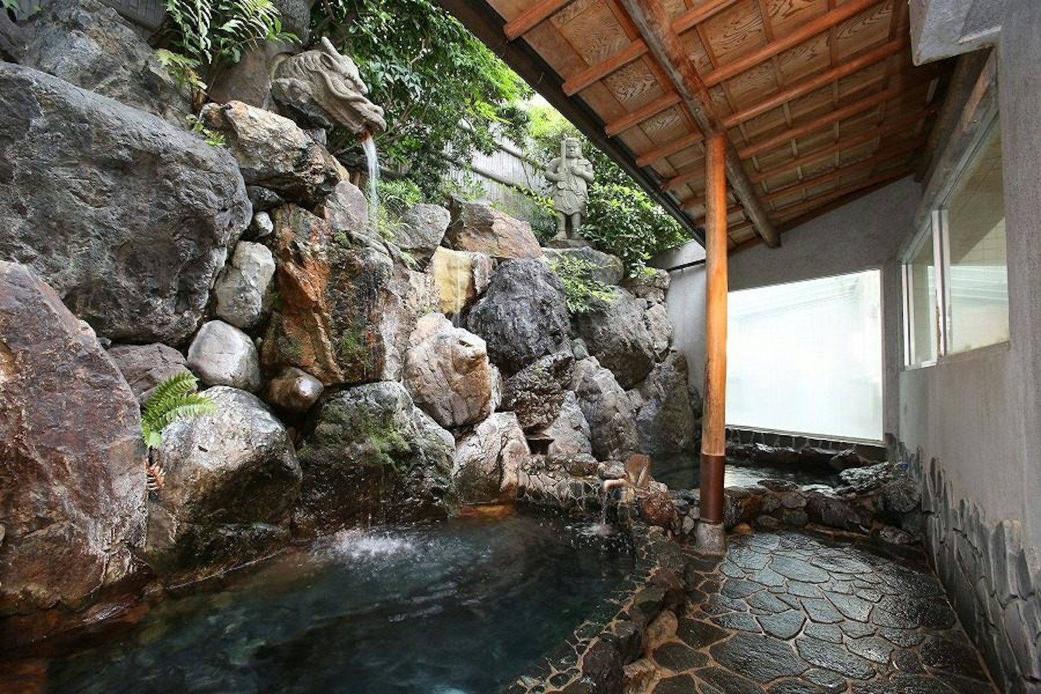 Funaoka Onsen A serene outdoor hot spring bath (onsen) with natural rock formations and a wooden roof. The water gently flows over the rocks, creating a tranquil atmosphere. Dense greenery surrounds the area, enhancing the peaceful, natural ambiance.