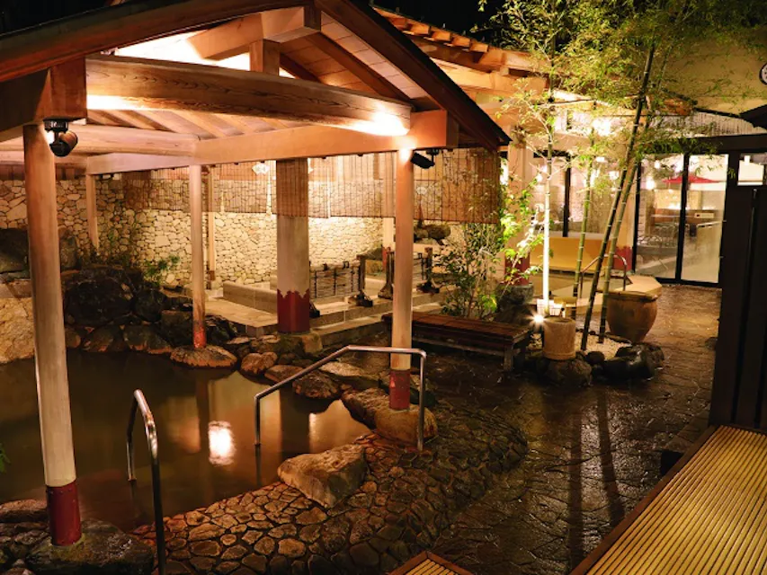 이슬녘의 일본 야외 온천 욕탕(온천)으로, 아늑한 조명, 나무 덮개, 돌길 및 대나무 식물 등 무성한 초목이 특징입니다. 물이 바위에 둘러싸인 목욕 공간에서 부드럽게 흐르며, 고요하고 매력적인 분위기를 자아냅니다.