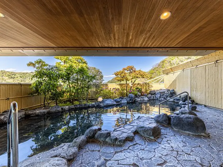 이 이미지는 맑은 하늘 아래 울창한 녹색 나무들로 둘러싸인 고요한 정원을 배경으로 한 야외 일본 온천(온천수)을 보여줍니다. 온천은 바위로 둘러싸인 가장자리를 가지고 있으며 주변을 반사하는 물이 있는 모든 것은 나무 지붕 아래에 있습니다.