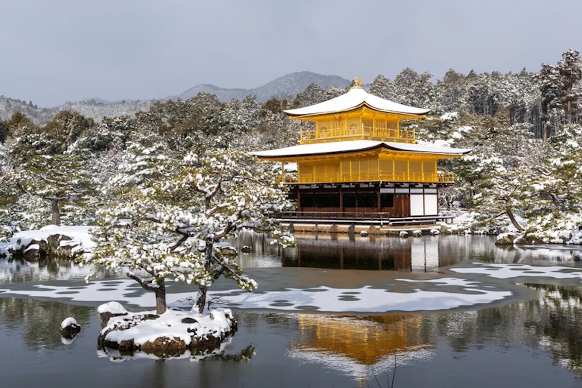 Snowy Kinkaku-ji Temple in winter A serene winter scene featuring Kinkaku-ji, the Golden Pavilion, in Kyoto, Japan. The temple is reflected in the calm pond surrounded by snow-covered trees and landscape. Hills faintly visible in the background under a cloudy sky.