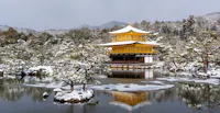 A serene winter scene featuring Kinkaku-ji, the Golden Pavilion, in Kyoto, Japan. The temple is reflected in the calm pond surrounded by snow-covered trees and landscape. Hills faintly visible in the background under a cloudy sky.
