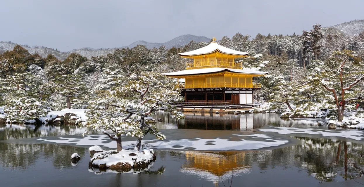 A serene winter scene featuring Kinkaku-ji, the Golden Pavilion, in Kyoto, Japan. The temple is reflected in the calm pond surrounded by snow-covered trees and landscape. Hills faintly visible in the background under a cloudy sky.