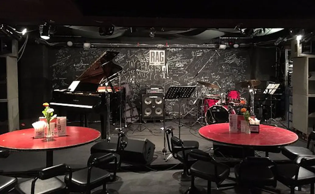 A dimly lit jazz club stage with a grand piano on the left, a drum set on the right, and microphones in the center. There are circular red tables with flowers and menus placed in the foreground, and a blackboard wall with various handwritten notes and doodles in the background.