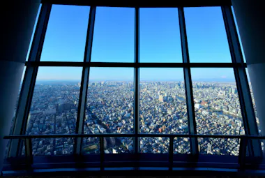 A panoramic cityscape view of Tokyo, Japan, seen through large floor-to-ceiling windows from a high observation deck on a clear, sunny day.