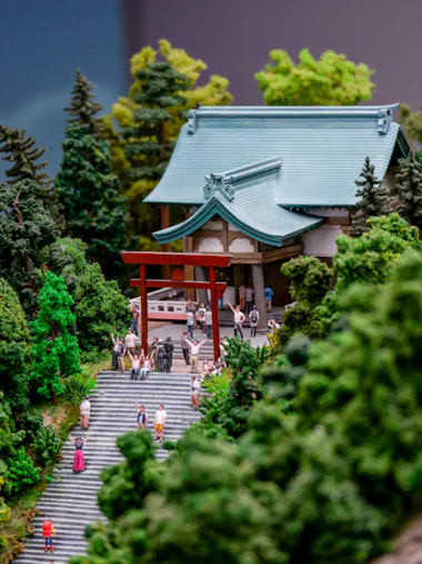 A miniature model of a Japanese shrine with a torii gate, stone steps, and small figurines of people surrounded by lush green trees.