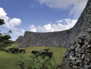 Ancient stone walls curve along a grassy landscape under a partly cloudy blue sky, with small palm trees and green foliage in the foreground.