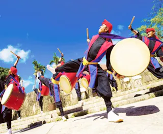 A group of performers dressed in traditional attire play large drums and raise their drumsticks, dancing energetically outdoors on stone steps under a bright blue sky.
