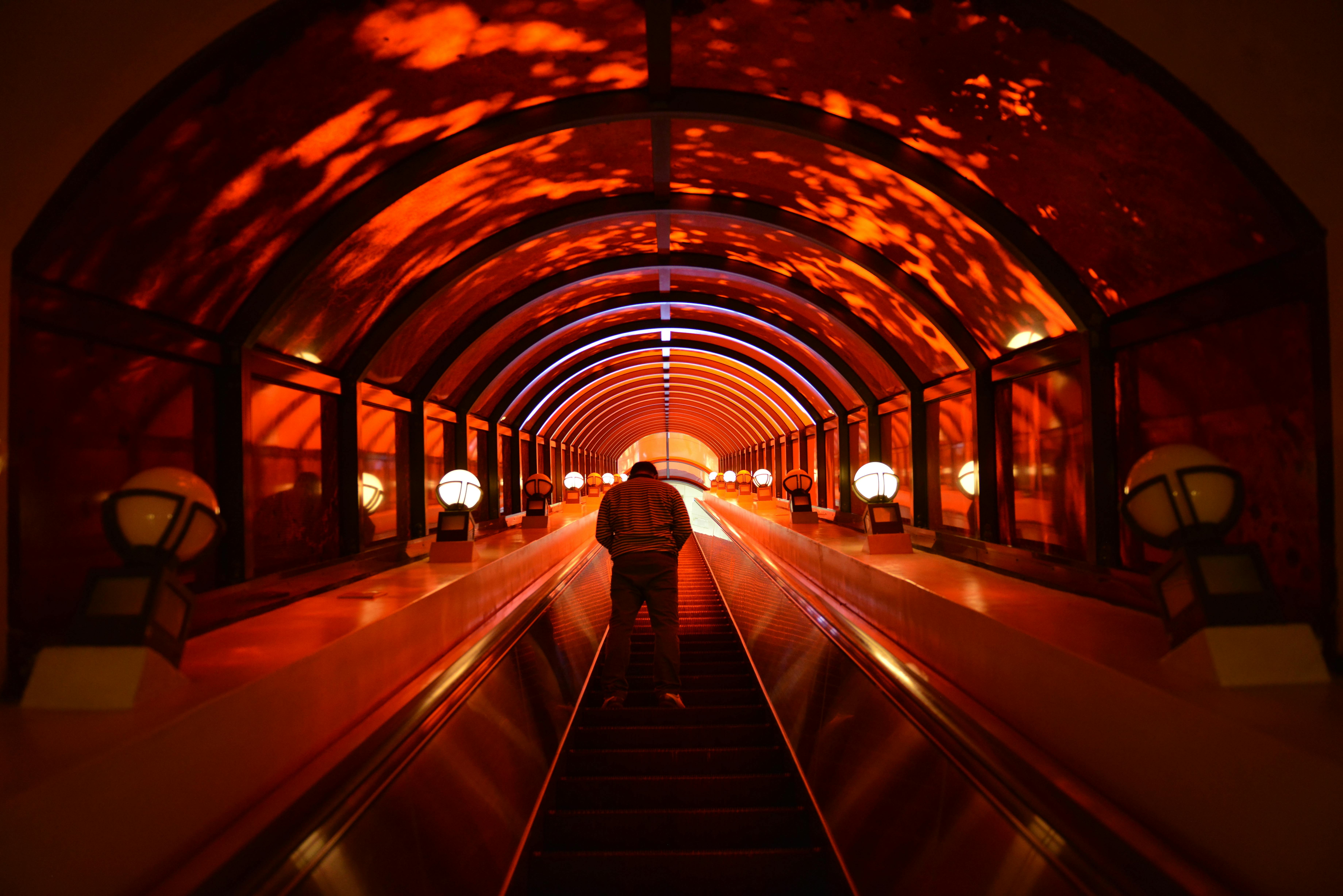 A person rides up a long escalator inside a tunnel illuminated with red and orange lights, creating a dramatic, glowing atmosphere. The ceiling is arched with reflections and lined with round lamps.