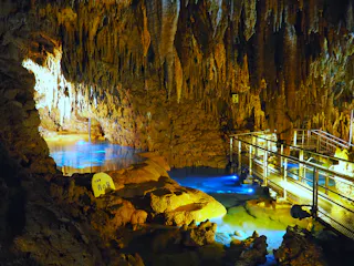 An illuminated limestone cave with stalactites hanging from the ceiling, a clear blue pool of water, a metal walkway with railings, and a stone marker with Japanese writing. The scene is warmly lit, creating a mystical atmosphere.