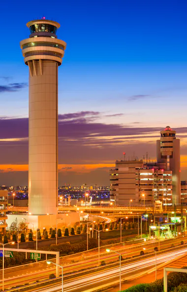 Haneda Airport A brightly lit air traffic control tower stands tall at an airport during sunset, surrounded by terminal buildings, roads, and parked cars, with a vibrant orange and blue sky in the background.
