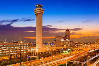 A brightly lit air traffic control tower stands tall at an airport during sunset, surrounded by terminal buildings, roads, and parked cars, with a vibrant orange and blue sky in the background.