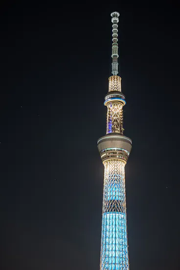 The Tokyo Skytree illuminated at night, glowing with blue and white lights against a dark sky, highlighting its tall, modern structure and intricate design.