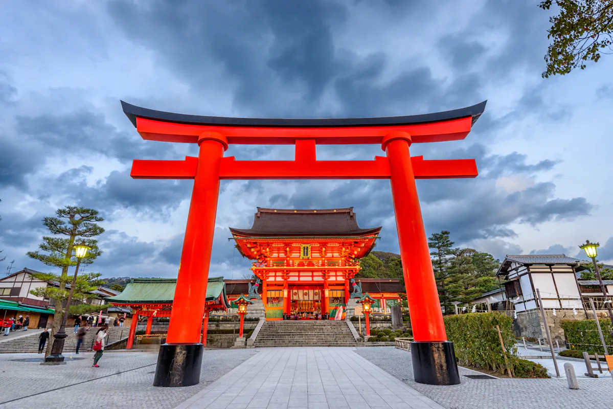 A stunning photo of the Fushimi Inari Shrine in Kyoto, Japan, features the iconic large red torii gate at the entrance. The gate stands tall against an overcast sky, with the shrine's traditional architecture visible in the background. Visitors explore the sacred site.