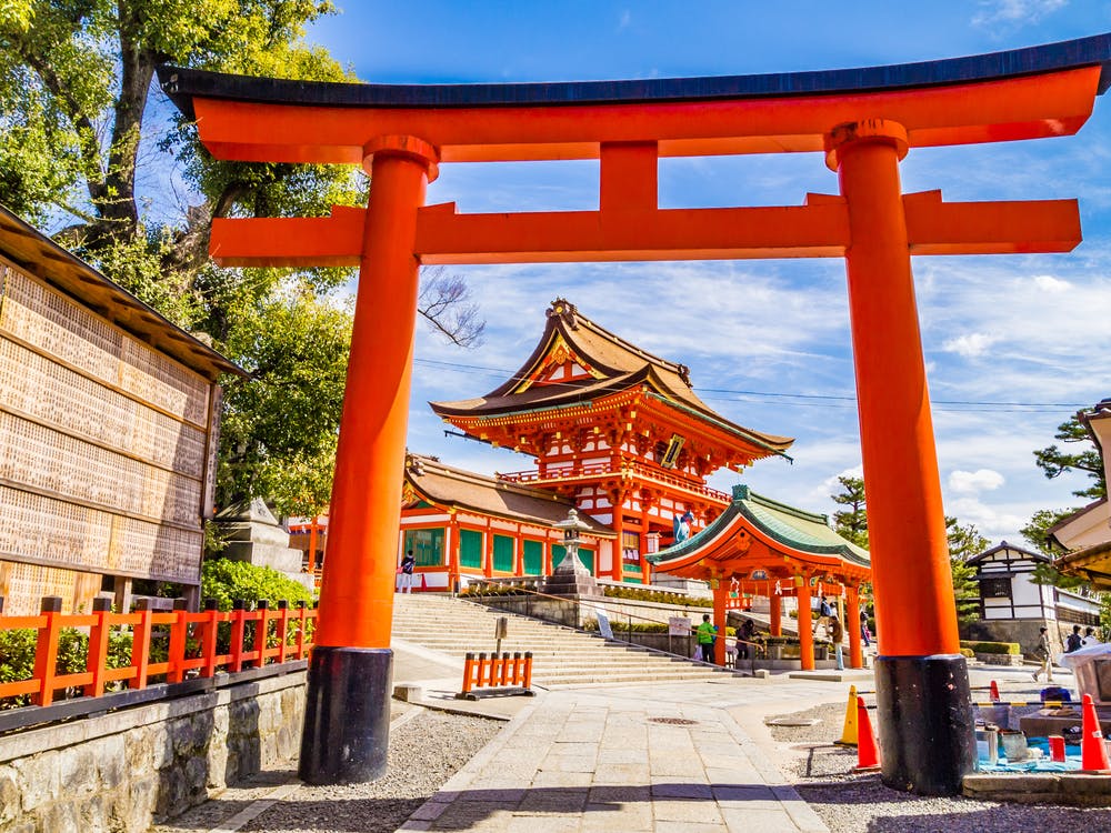 A large red torii gate stands at the entrance to Fushimi Inari Shrine in Kyoto, Japan. Beyond the gate are traditional Japanese buildings featuring ornate wooden architecture and vibrant red accents. The sky is clear and blue, and trees surround the shrine.