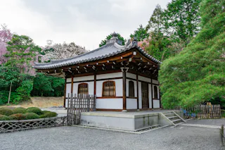 A traditional Japanese building with a tiled roof stands among trees and bushes. It has wooden beams and white walls with barred windows. A small set of steps leads to the entrance, and gravel covers the foreground. The sky is partly cloudy.