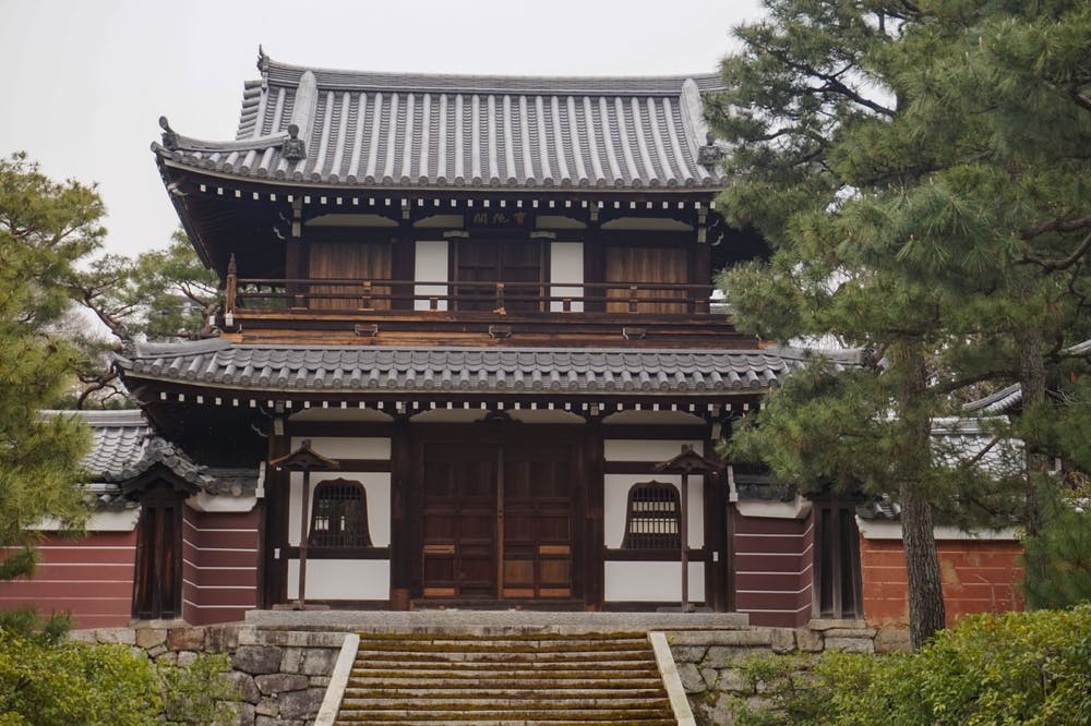 A traditional Japanese wooden building with a tiled roof, surrounded by greenery. The structure has two stories and features elements of classic Japanese architecture. Stairs lead up to the entrance, which has wooden doors and lattice windows.