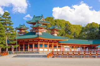 A traditional Japanese shrine with green-tiled roofs and vibrant orange wooden structures set against the backdrop of lush green trees and a blue sky with scattered clouds. The intricate architecture showcases multi-tiered roofs and ornate carvings.
