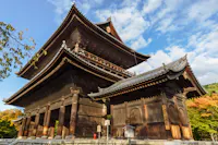 A traditional wooden Japanese temple with intricately designed eaves and multiple tiers, set against a backdrop of a partly cloudy blue sky. The temple is surrounded by greenery, with some trees displaying autumn colors.