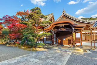 A traditional Japanese temple with ornate wooden architecture, featuring a curved roof and intricate carvings, set against a backdrop of lush greenery and autumn foliage with vibrant red and orange leaves. The sky is clear and blue.