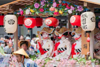 A group of people dressed in traditional Japanese kimonos and straw hats participate in a festival parade. They are on a decorated float adorned with colorful flowers and red lanterns with kanji characters. The background shows a crowd of onlookers.