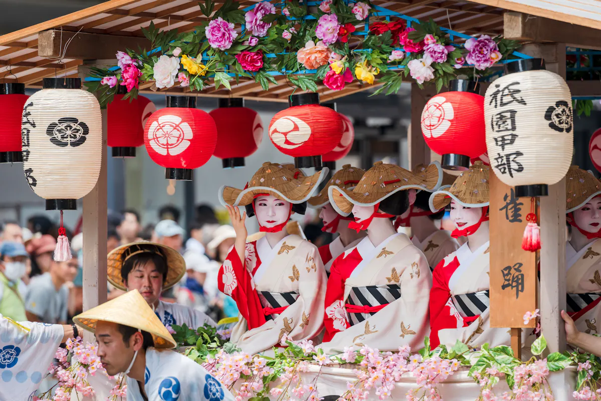 A group of people dressed in traditional Japanese kimonos and straw hats participate in a festival parade. They are on a decorated float adorned with colorful flowers and red lanterns with kanji characters. The background shows a crowd of onlookers.