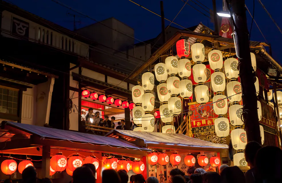 A bustling night scene of a traditional Japanese festival with numerous glowing paper lanterns hanging from wooden structures. People gather around, enjoying the evening ambiance as lanterns illuminate the area with a warm, festive glow.