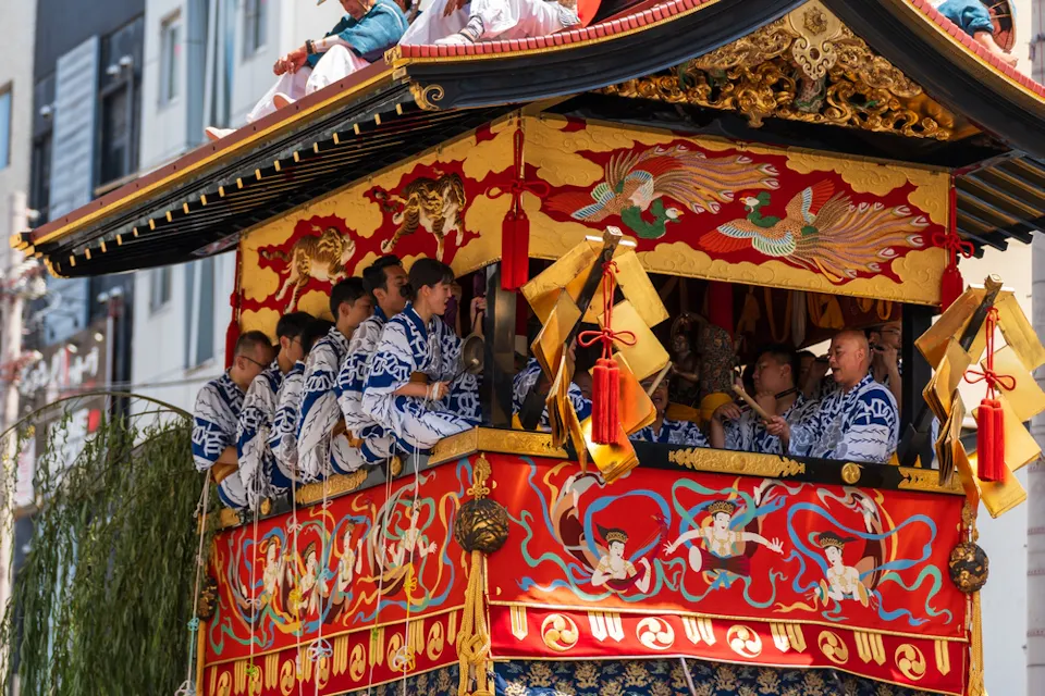 A group of men wearing traditional blue and white garments stand on a colorful, highly decorated float featuring red, gold, and intricate designs. The float is adorned with hanging ornaments and lush greenery, participating in a festive parade.