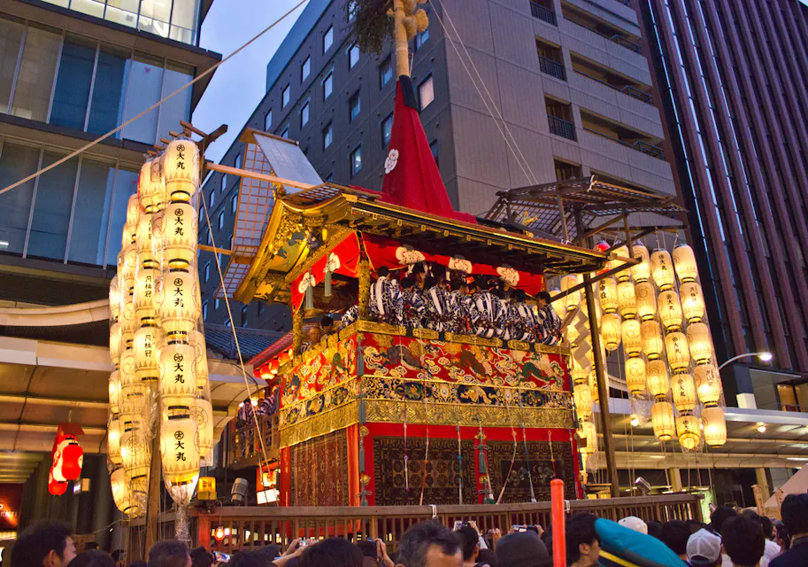 A traditional Japanese float adorned with intricate decorations and red fabric is paraded through a city street. The float is illuminated by numerous paper lanterns. A crowd of people surrounds the float, and tall buildings are visible in the background.