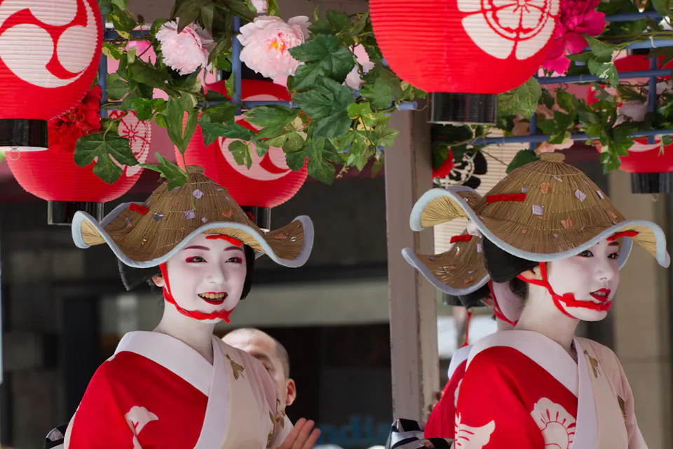 Two women wearing traditional Japanese makeup and attire, with white faces and red and black details, are dressed in red and white kimonos. They are under red lanterns decorated with floral patterns and are wearing wide-brimmed hats adorned with butterflies.