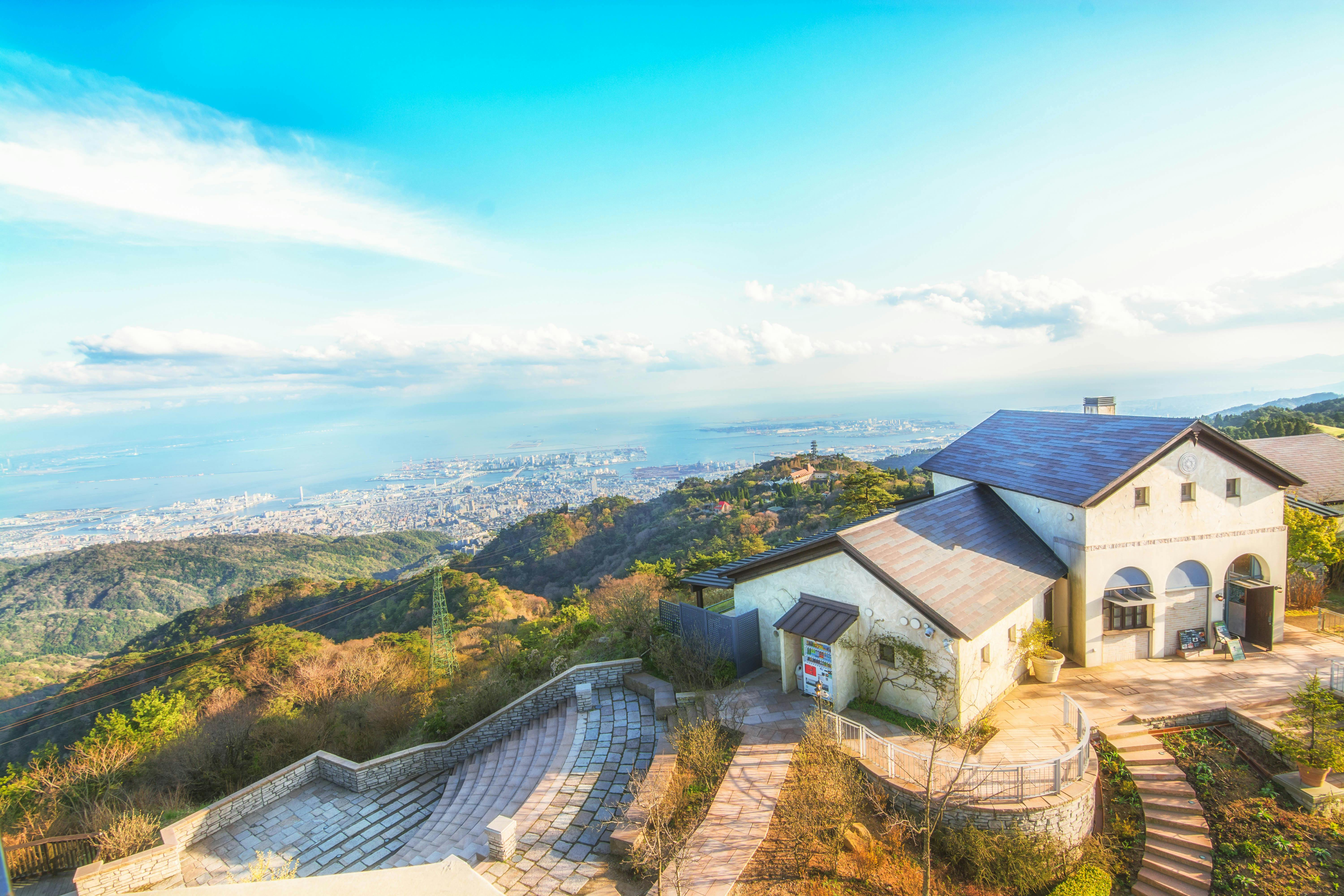 A scenic view of a white building with arched windows on a hillside, overlooking a city and coast far below, with blue sky and scattered clouds above.