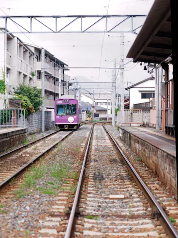 A purple tram approaches a small, quiet train station lined with buildings and greenery on a cloudy day. The tracks are flanked by concrete platforms and power lines stretch overhead.