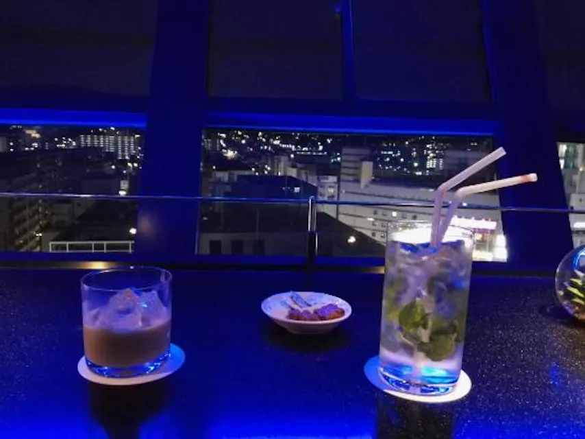 Sky Lounge Kuu A dimly lit rooftop bar at night, showcasing a cityscape through large windows. Two drinks, one in a highball glass with mint leaves and two straws, and another in a tumbler, rest on a table alongside a small plate of snacks. Blue lighting creates a serene ambiance.