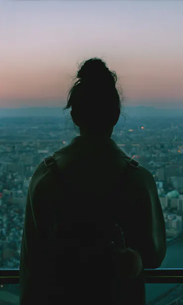 Tokyo Skytree A person with their hair in a bun stands indoors, facing large windows and looking out over a sprawling cityscape at dusk, with soft pink and blue hues in the sky.