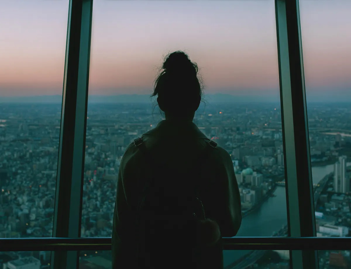 Tokyo Skytree A person with their hair in a bun stands indoors, facing large windows and looking out over a sprawling cityscape at dusk, with soft pink and blue hues in the sky.