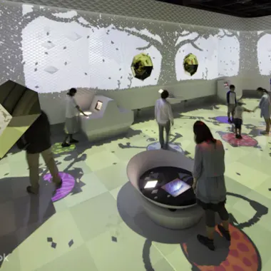 Miraikan Museum People interact with digital touchscreens and walk in a bright, immersive art exhibit with colorful, animated projections covering the walls and floor, creating a futuristic and playful environment.