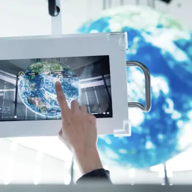 Miraikan Museum A person’s hand interacts with a digital touchscreen displaying a map of Earth, while a large globe hangs in the blurred background, suggesting a high-tech, futuristic environment.