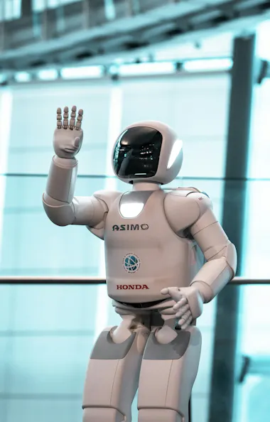 Miraikan Museum A white humanoid robot labeled "ASIMO" and "Honda" stands indoors, waving its right hand in front of a large glass window with a blurred, modern background.
