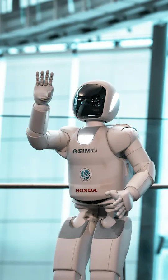 Miraikan Museum A white humanoid robot labeled "ASIMO" and "Honda" stands indoors, waving its right hand in front of a large glass window with a blurred, modern background.