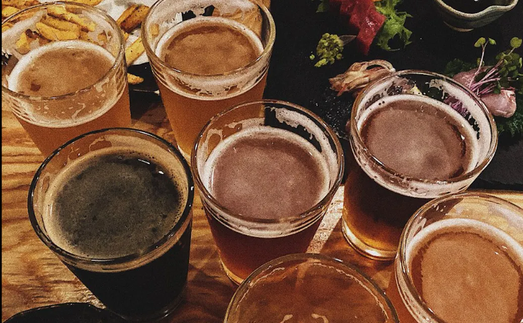 Beer Komachi A variety of beer glasses filled with different types of beer are arranged on a wooden surface. In the background, there are plates with food items, including some fries and a partially visible dish with greens.